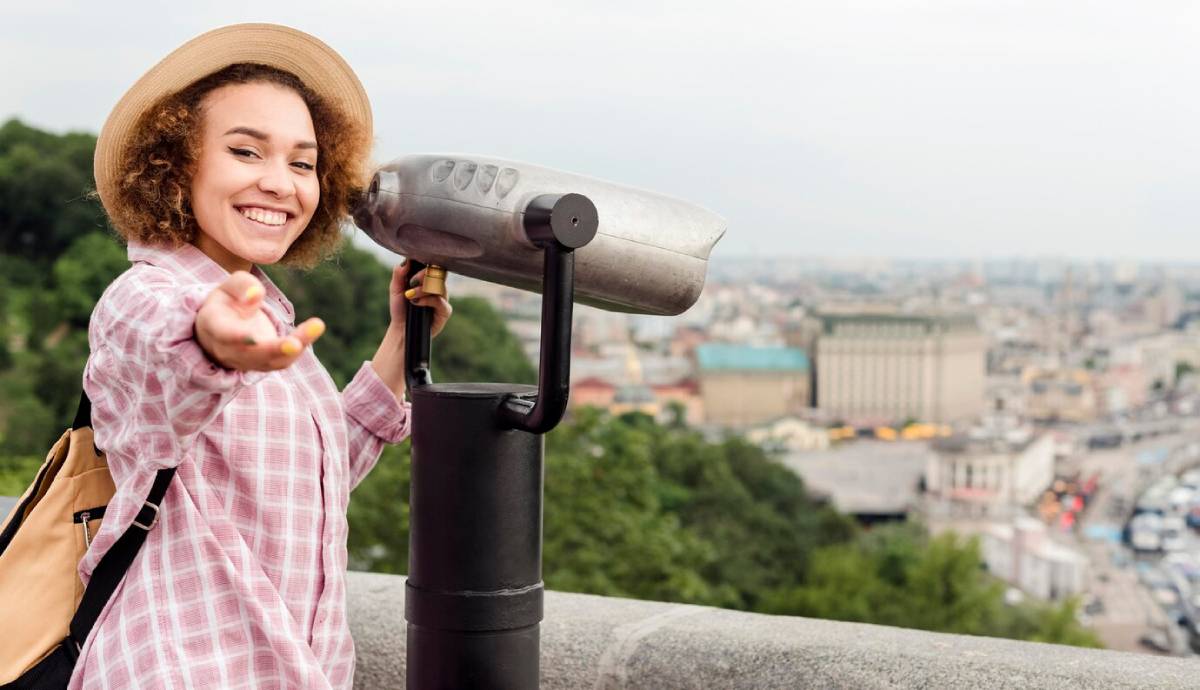 Young woman using a viewing telescope on a city overlook, smiling and reaching out. 
