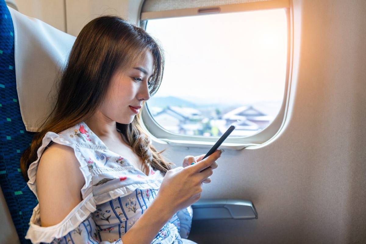 Young woman using smartphone on airplane by window, casual attire. 