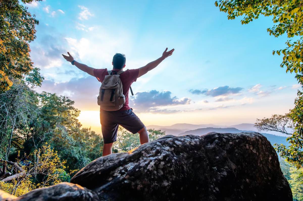 Man with arms raised in triumph on a mountain top at sunset, surrounded by trees and overlooking distant hills. 