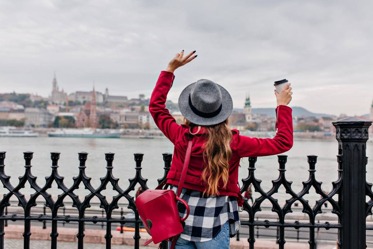 Woman in a red jacket and gray hat holding a coffee cup, greeting a riverfront cityscape from a wrought iron fence.