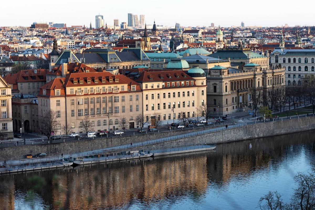 Aerial view of Prague with historic buildings along the riverfront, reflecting in the calm water of the Vltava River.