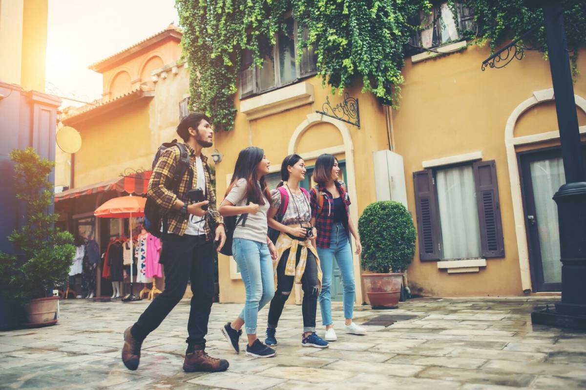  Four young adults with backpacks exploring and walking through a picturesque European-style street, exhibiting curiosity and enjoyment.