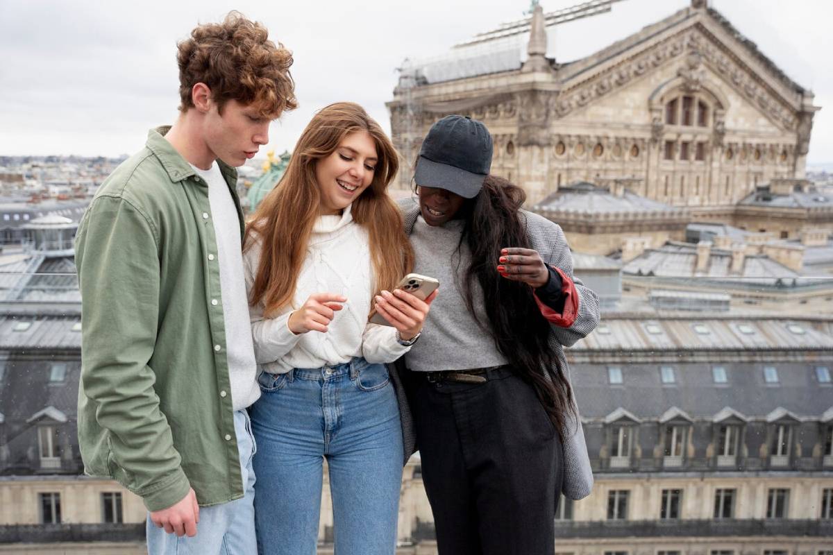 Three young friends looking at a smartphone together against a backdrop of historic city buildings. 