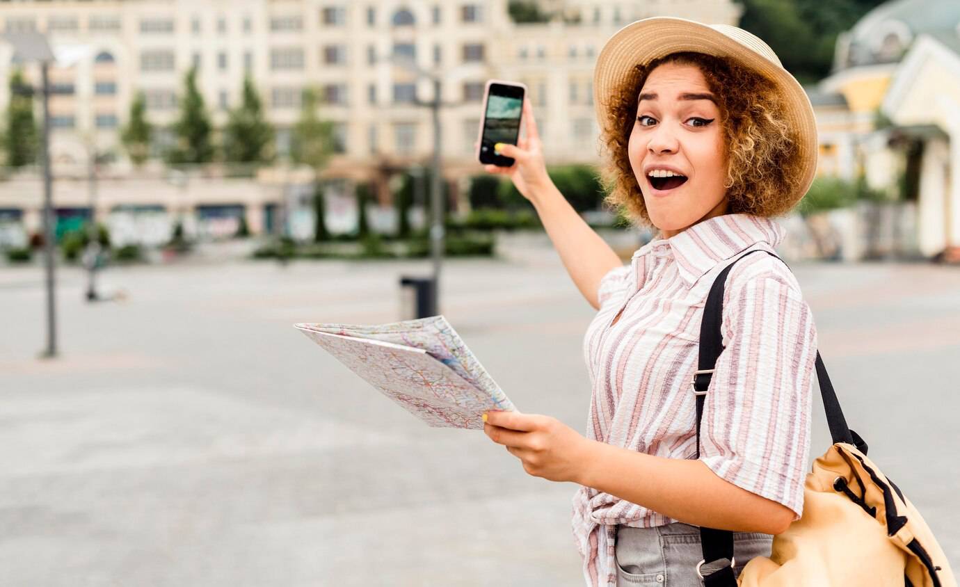 A joyful young woman with a hat shows her phone and holds a map in a lively city square.